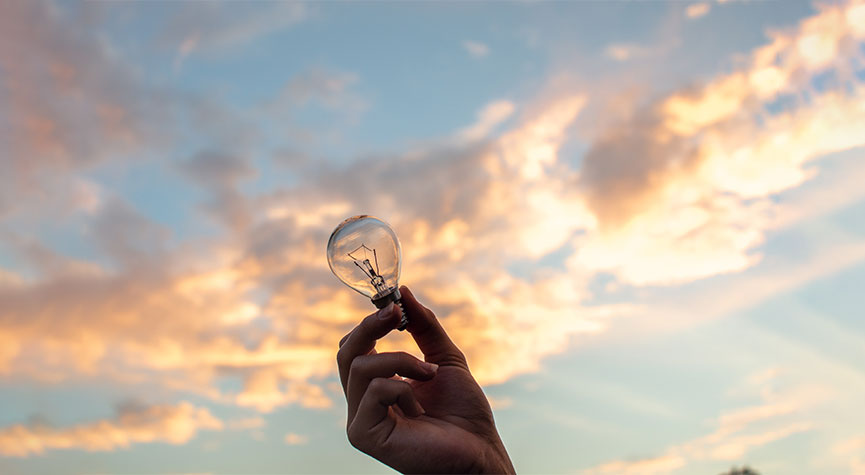 a person outside, only their hand showing and they're holding a lightbulb with a beautiful sky sunsetting background