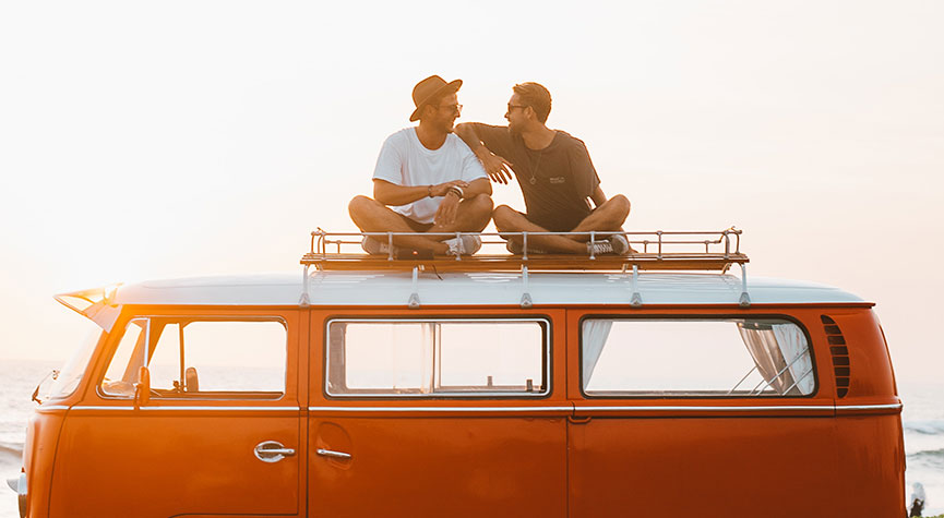 two men sitting on top of a orange volkswagan van talking and smiling