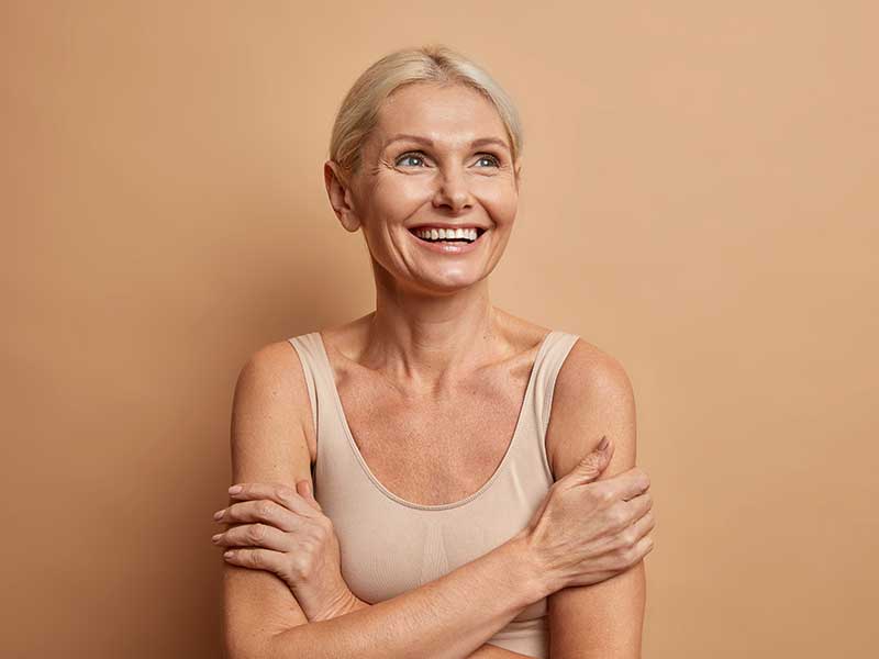 a woman over 60 years old hugging herself and smiling in front of a tan background