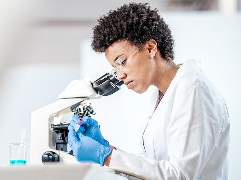 a woman in a laboratory wearing blue gloves and a white lab coat looking through a microscope