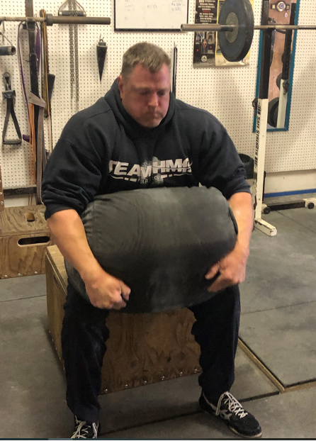 Renowned Hall of Fame powerlifter Brad Gillingham demonstrates exceptional strength and technique as he flawlessly executes a sandbag clean and box squat during a training session. The image showcases the precision and power inherent in elite powerlifting performance.